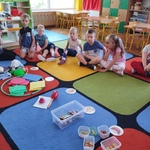 Dzieci siedzą w kole. Na dywanie leżą różne przedmioty i obrazki narządó zmysłów .jpg